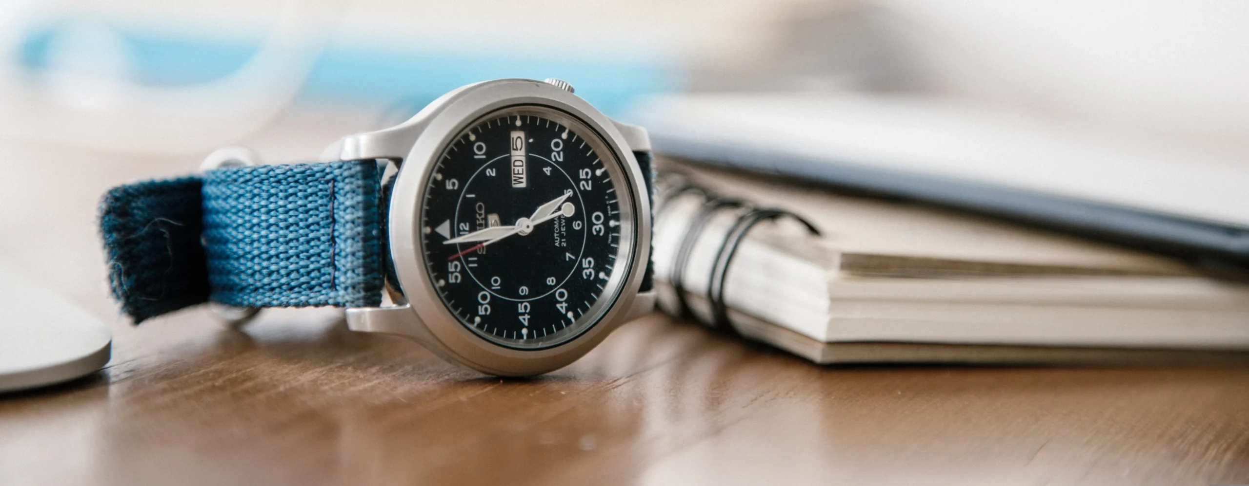 A close-up of a Seiko 5 Sports watch with a blue strap resting on a wooden desk next to a notebook.